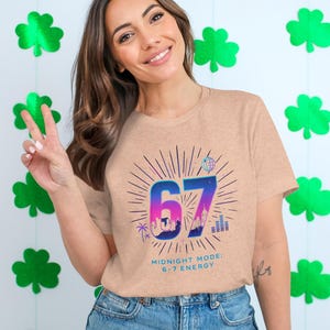 May include: A light peach-colored t-shirt with a graphic design featuring the number 67 in blue and purple, a cityscape silhouette, and the text "MIDNIGHT MODE: 6-7 ENERGY". The background includes green shamrock decorations.