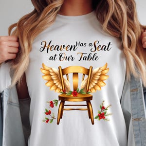 May include: White t-shirt with a golden chair design, angel wings, and the text "Heaven Has a Seat at Our Table." The chair is decorated with red berries and foliage. The graphic is centrally placed on the shirt.