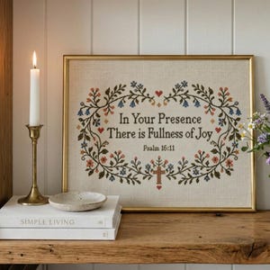 May include: A framed cross-stitch embroidery on light beige fabric featuring a floral wreath surrounding the text In Your Presence There is Fullness of Joy Psalm 16:11 with a small cross at the bottom, displayed in a gold-toned frame.