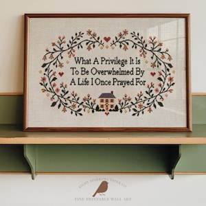 May include: A framed cross-stitch embroidery on light fabric featuring a floral wreath border with small red hearts and a house at the bottom. The text reads: What A Privilege It Is To Be Overwhelmed By A Life I Once Prayed For. The frame is wood.