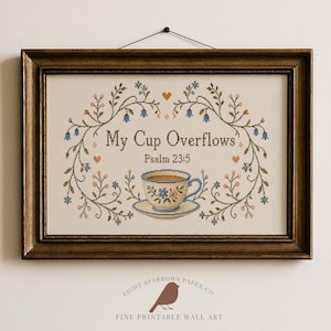 Könnte beinhalten: Eine gerahmte Kreuzstichstickerei mit dem Text My Cup Overflows und Psalm 23:5. Das Design zeigt eine zentrale Teetasse auf einer Untertasse, umgeben von einem Blumenkranz mit blauen und rosa Blüten, grünen Blättern und kleinen orangefarbenen Herzen.