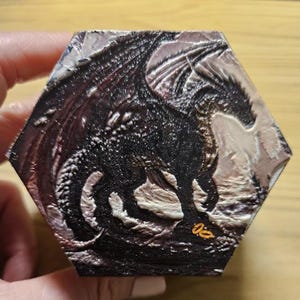 May include: A hexagonal tile featuring a textured, dark relief carving of a dragon with wings spread. The dragon is rendered in shades of black and metallic silver, with a small, bright orange symbol visible near the bottom right corner.
