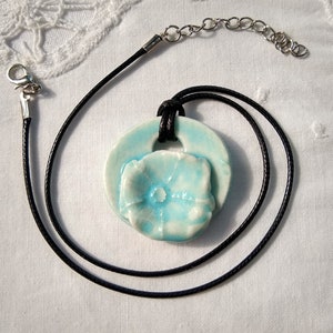 May include: A ceramic pendant necklace featuring a light blue flower design. The pendant is attached to a black cord with a silver-tone chain and clasp. The necklace is displayed on a white, textured surface.