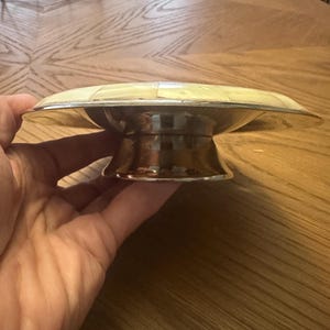 May include: A circular pedestal dish featuring a metallic silver-toned base and a shallow bowl inlaid with a mosaic of cream-colored, iridescent shell tiles arranged in a geometric pattern, creating a smooth, reflective surface on the top.