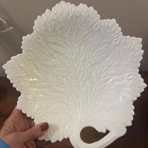 May include: A white, leaf-shaped serving platter with intricate, raised vein detailing. The platter has a curved handle and a scalloped edge. The design is reminiscent of a large, stylized leaf, perfect for serving appetizers or desserts.