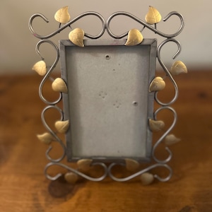 May include: A rectangular metal picture frame with a silver finish and gold-colored leaf accents. The frame has a decorative, swirling design with small, gold-colored leaves. The frame is designed to hold a photograph.