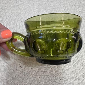 May include: Green glass teacup with a handle. The cup has a rounded body with embossed circular designs and a decorative rim. The handle is curved and attached to the side of the cup. The glass has a slight tint.