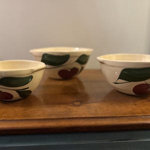 May include: Three ceramic bowls with a cream-colored base and a cherry design. Each bowl features a red cherry and green leaves. The bowls are arranged on a wooden surface.
