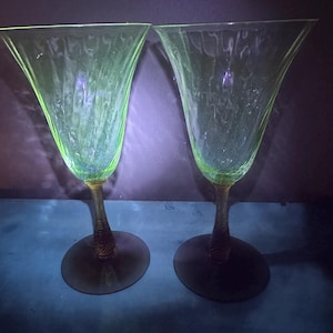 May include: Two vintage green-tinted glass goblets. The bowls are flared and textured with vertical lines. The stems are twisted, and the bases are dark green. The glasses are set against a dark background.