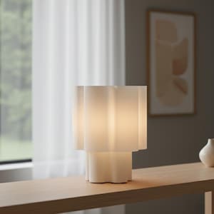 Könnte beinhalten: Eine moderne, weiße Tischlampe mit einem einzigartigen, geschichteten Design. Die Lampe hat einen wellenförmigen, gewellten Schirm und Sockel und strahlt ein warmes, weiches Licht aus. Die Lampe steht auf einem hellen Holztisch und schafft eine minimalistische Ästhetik.
