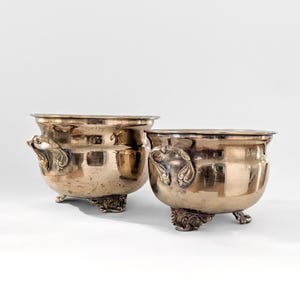 May include: Two antique silver-toned metal bowls. Each bowl has a rounded body, ornate handles, and decorative feet. The bowls have a tarnished appearance, suggesting age and use. The background is plain white.