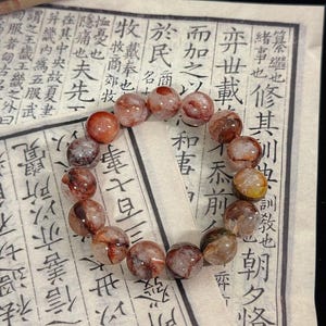 May include: A bracelet made of spherical beads in shades of reddish-brown and white. The beads are translucent and have internal inclusions. The bracelet is displayed on a background of aged paper with Asian script.