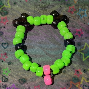 Può includere: Un braccialetto di perline verde neon e nero con un ciondolo rosa. Il braccialetto è composto da piccole perline rotonde infilate insieme. Il ciondolo è composto da due perline rettangolari rosa. Lo sfondo è un motivo colorato.