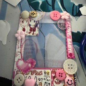 May include: A decorative photo frame with a clear center, adorned with various embellishments. It features pink, white, and gold buttons, a pink heart, and small flower accents. The frame includes printed images and text, with the words "LOVE, BABY!" visible.