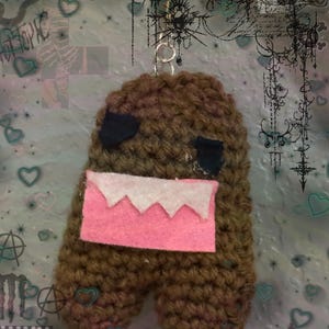 May include: A handmade brown crochet Domo-kun keychain. The keychain features black eyes, a pink mouth with white teeth, and a silver keyring. The background has a collage of hearts, stars, and text.