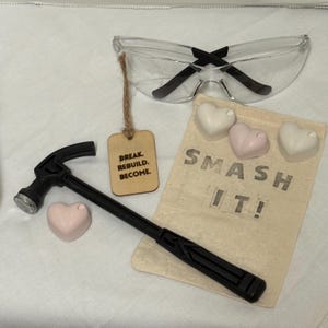May include: A black hammer, safety glasses, and a small canvas bag with the words "SMASH IT!" printed on it. The bag contains three heart-shaped objects. A wooden tag reads "BREAK. REBUILD. BECOME."