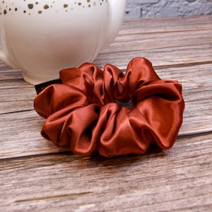 May include: A rust-colored satin scrunchie sits on a wooden surface, with a white teapot with gold accents in the background. The scrunchie is gathered and has a shiny, textured appearance. This hair accessory adds a touch of elegance.