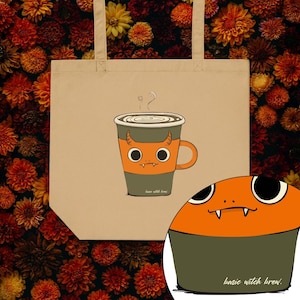 Könnte beinhalten: Beige Tragetasche mit einem Cartoon-Kaffeebecher-Design. Der Becher ist orange und grau mit Teufelshörnern und Augen. Der Text "basic witch brew" befindet sich am unteren Rand des Bechers und in der unteren rechten Ecke. Dampf steigt aus der Tasse auf.
