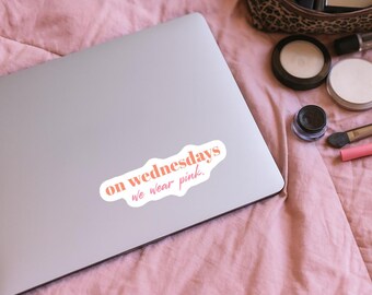 May include: A white sticker with the text "on Wednesdays we wear pink" in a pink and orange font, placed on a silver laptop. The laptop is on a pink surface, with makeup items in the background.