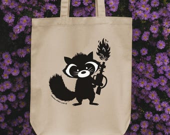 May include: Beige tote bag with a black and white raccoon illustration holding a flamethrower. The raccoon has a white face and a fluffy tail. The text "random chaos club" is printed at the bottom. The bag has two handles.