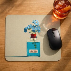 May include: A beige mousepad with a blue-tinted glass bottle containing blue liquid and a sprig of greenery. The bottle holds a small bouquet of blue flowers. A black wireless mouse and a glass of amber liquid with ice and an orange slice are also on the wooden surface.