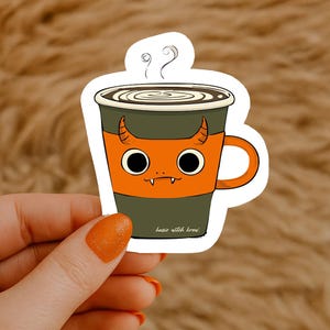 Könnte beinhalten: Ein Aufkleber mit einer Cartoon-Kaffeetasse mit einem Teufelsgesicht. Die Tasse ist orange und grün, mit aufsteigendem Dampf. Der Text "basic witch brew" befindet sich unten.