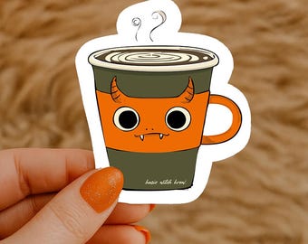 Op de afbeelding: Een sticker met een cartoon koffiekopje met een duivelsgezicht. De beker is oranje en groen, met stoom die opstijgt. De tekst "basic witch brew" staat onderaan.