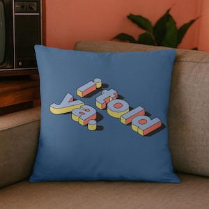 Könnte beinhalten: Ein blaues Zierkissen mit dem Schriftzug "Ya told!" in einem 3D-Schriftstil im Retro-Look. Der Text ist in Gelb, Rot und Blau gehalten und hat einen Schatteneffekt. Das Kissen liegt auf einem grauen Sofa.