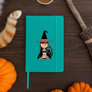 Könnte beinhalten: Türkisfarbenes Notizbuch mit einer Cartoon-Hexe und einer schwarzen Katze. Die Hexe trägt einen schwarzen Hut und hält eine Kaffeetasse. Das Notizbuch liegt auf einer dunkelbraunen Holzoberfläche, mit einem Kürbis und einem Besen im Hintergrund.
