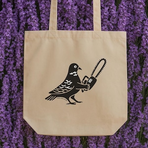 Könnte beinhalten: Beige Tragetasche mit einer schwarzen Illustration einer Taube, die eine Kettensäge hält. Die Taube hat detaillierte Flügel- und Schwanzfedern. Die Tasche hat zwei Griffe und ist aus robustem Stoff gefertigt. Der Hintergrund ist ein Feld aus lila Blumen.