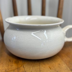 May include: Cream-colored, antique chamber pot with a rounded body and a single handle. The ceramic pot has a wide rim and a crackled glaze, suggesting age and use. It sits on a wooden chair, showcasing its vintage aesthetic.