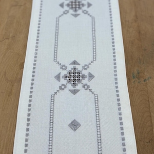 May include: A rectangular white table runner with a gray geometric design. The design includes diamond, square, and rectangular shapes. The runner is made of a woven fabric and is displayed on a wooden surface.