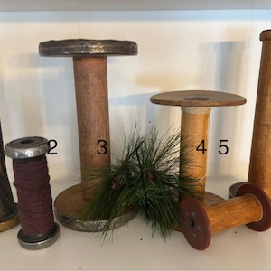 May include: A collection of vintage wooden spools in various sizes and shapes. The spools are made of wood and metal, with some featuring a dark, aged appearance. A sprig of green pine needles adds a touch of natural decor.