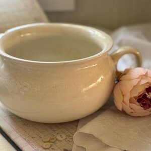 May include: A cream-colored ceramic chamber pot with a rounded body, a wide rim, and a single side handle. The surface features a visible crackle glaze finish throughout. A soft pink artificial peony flower rests beside the handle.
