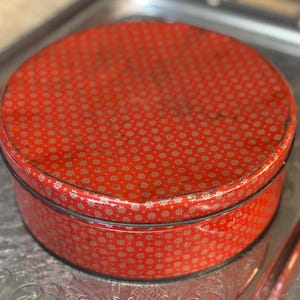 May include: A round, vintage red tin with a lid. The tin is covered in a repeating pattern of small, gold-colored starbursts. The tin sits on a silver tray. The tin has a black rim.