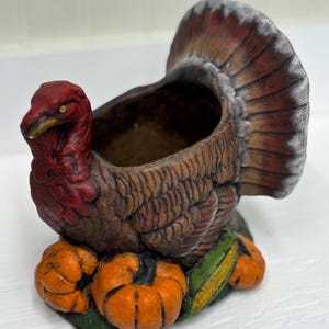 May include: A ceramic turkey planter with a red head and brown body, perched atop orange pumpkins and green leaves. The turkey's tail feathers are a mix of brown and gray. A decorative item for fall or Thanksgiving.