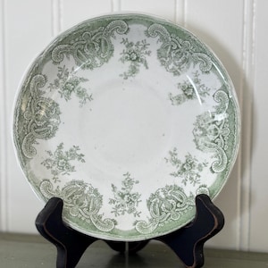 May include: A white ceramic plate with a green floral and paisley design. The plate is displayed on a black wooden stand. The plate's rim has a scalloped edge. The floral pattern is repeated around the rim and in the center of the plate.