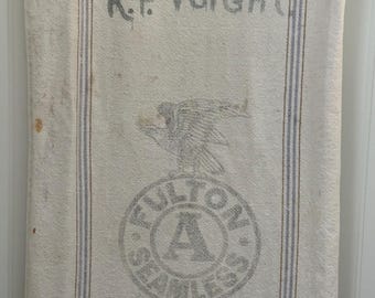 May include: Cream-colored vintage textile with blue and brown stripes, featuring the name "R.F. Voight" and a circular logo with an eagle and the words "Fulton Seamless Extra Heavy." The textile is held by a wooden bar.