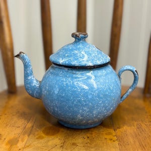 May include: A light blue enamel teapot featuring a white speckled pattern throughout the body and lid. The teapot has a curved spout, a rounded handle, and a dark brown rim along the lid and opening, showing signs of wear on the edges.