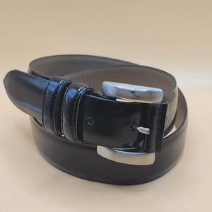 Puede incluir: Un cinturón de cuero negro con un acabado brillante y una hebilla de color plateado. El cinturón está enrollado, mostrando el interior del cinturón. La hebilla es rectangular y tiene un acabado plateado.
