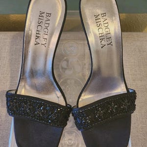 Puede incluir: Un par de sandalias de tacón Badgley Mischka de satén negro. Las sandalias tienen punta cuadrada y una tira en la parte superior del pie adornada con cuentas negras. La plantilla es plateada con el logotipo de Badgley Mischka.
