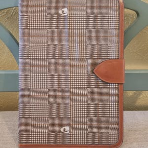 May include: A rectangular document portfolio featuring a brown and white houndstooth pattern with thin tan grid lines. The case is bordered by smooth brown leather trim and includes a matching leather tab closure with a small embossed logo emblem.