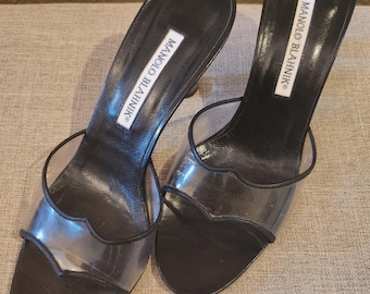 May include: A pair of black slide sandals featuring clear transparent PVC straps with a scalloped edge design. The footbed is black leather with a white rectangular label displaying the text MANOLO BLAHNIK in black capital letters.