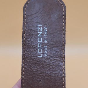 May include: Close-up of a brown leather belt with the text "LORENZI MADE IN ITALY" in white. The belt has a textured surface and visible stitching along the edges. The leather shows signs of wear.