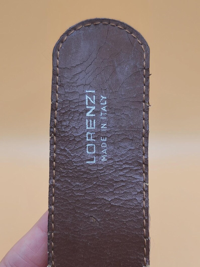 May include: Close-up of a brown leather belt with the text "LORENZI MADE IN ITALY" in white. The belt has a textured surface and visible stitching along the edges. The leather shows signs of wear.