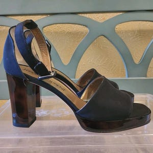 May include: Black platform sandals with a high heel and ankle strap. The sandals have a suede upper and a glossy platform sole. The heel is a dark tortoiseshell color. The brand name "Yves Saint Laurent" is visible on the side.
