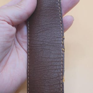 May include: Close-up of a brown leather belt with visible cracking and wear. The belt has a textured surface and is stitched with a lighter brown thread along the edges. The leather shows signs of age and use.