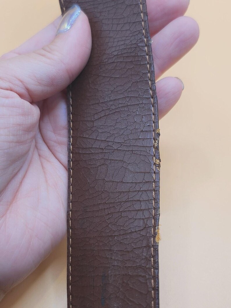May include: Close-up of a brown leather belt with visible cracking and wear. The belt has a textured surface and is stitched with a lighter brown thread along the edges. The leather shows signs of age and use.
