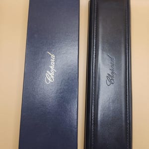 May include: A rectangular navy blue cardboard box and a long, slim black leather-textured case, both featuring the Chopard brand name printed in gold lettering on their top surfaces. The items are positioned side-by-side in a vertical orientation.