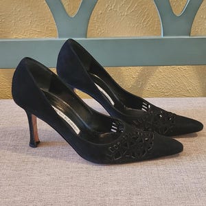 Puede incluir: Un par de tacones altos de ante negro con punta. Los tacones tienen un diseño calado en la parte delantera y un tacón de aguja. La marca "Manolo Blahnik" es visible en la plantilla.
